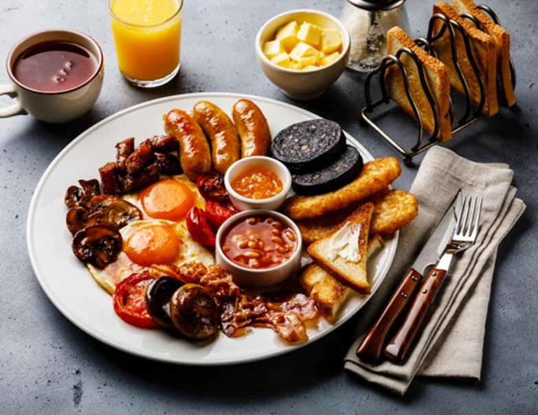 A large plate filled with a classic full English breakfast, including sausages, bacon, eggs, black pudding, hash browns, beans, tomatoes, mushrooms, and toast, accompanied by orange juice and tea