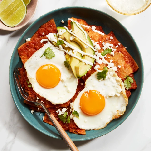 Red chilaquiles topped with two fried eggs, avocado slices, crumbled cheese, and cilantro, served on a blue plate.