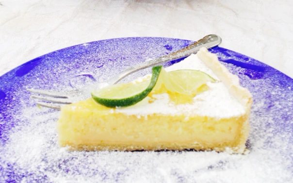 Slice of lemon tart on a blue plate with lime garnish.